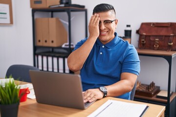Young hispanic man working at the office with laptop covering one eye with hand, confident smile on face and surprise emotion.