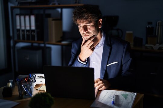 Hispanic young man working at the office at night looking confident at the camera with smile with crossed arms and hand raised on chin. thinking positive.