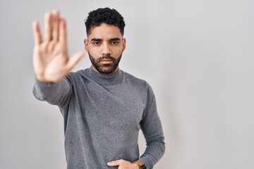 Hispanic man with beard standing over white background doing stop sing with palm of the hand. warning expression with negative and serious gesture on the face.