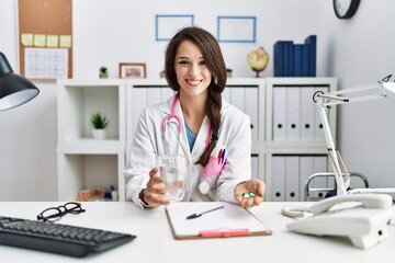 Young doctor woman holding glass of water and prescription pills smiling with a happy and cool smile on face. showing teeth.