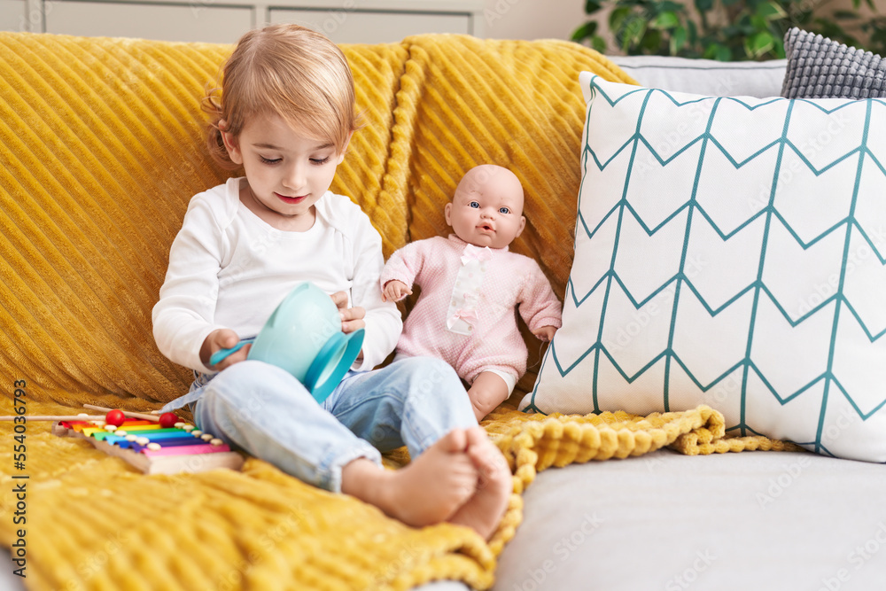 Poster Adorable caucasian girl playing with baby doll sitting on sofa at home