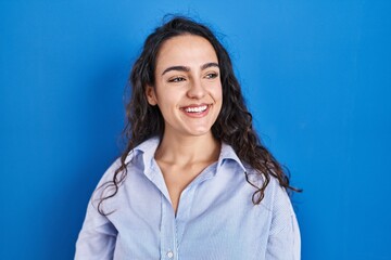 Young brunette woman standing over blue background looking away to side with smile on face, natural...