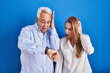Middle age hispanic couple standing over blue background looking at the watch time worried, afraid of getting late