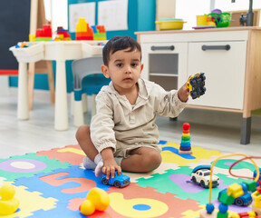 Adorable hispanic toddler playing with car toy sitting on floor at kindergarten