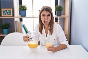 Young hispanic woman drinking glass of orange juice angry and mad screaming frustrated and furious, shouting with anger. rage and aggressive concept.