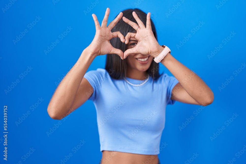 Sticker brunette young woman standing over blue background doing heart shape with hand and fingers smiling l