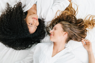 Two young beautiful smiling women with dark hair in white bathrobes lying on bed, High angle view of two charming cheerful girls in bedroom
