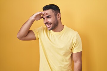 Fototapeta na wymiar Young hispanic man standing over yellow background very happy and smiling looking far away with hand over head. searching concept.