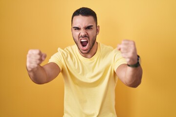 Fototapeta premium Young hispanic man standing over yellow background angry and mad raising fists frustrated and furious while shouting with anger. rage and aggressive concept.