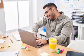 Young hispanic man business worker using laptop talking on smartphone at office