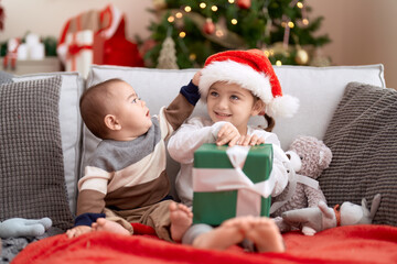 Fototapeta na wymiar Brother and sister opening gift sitting on sofa by christmas tree at home
