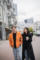 Smiling asian woman holding coffee to go and looking at boyfriend on urban street.