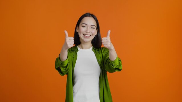 Positive young Asian woman shows thumbs-up smiles cheerfully
