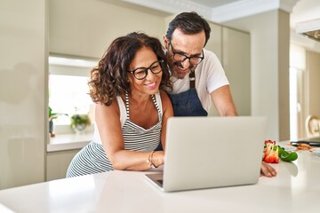 Middle age hispanic couple cooking and using laptop at kitchen