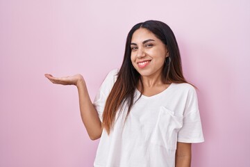 Young arab woman standing over pink background smiling cheerful presenting and pointing with palm of hand looking at the camera.