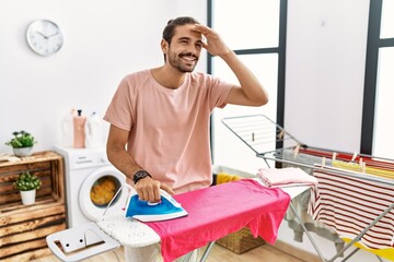 Young hispanic man ironing clothes at home very happy and smiling looking far away with hand over head. searching concept.