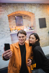 Smiling multiethnic couple taking selfie on cellphone in cafe.