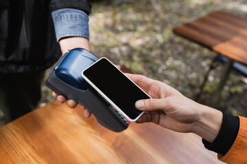 Cropped view of young man paying with smartphone near waitress with payment terminal in outdoor cafe.