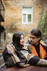 Young multiethnic couple in blanket looking at each other in outdoor cafe.