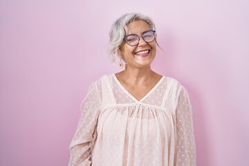 Middle age woman with grey hair standing over pink background winking looking at the camera with...