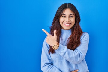Hispanic young woman standing over blue background cheerful with a smile on face pointing with hand and finger up to the side with happy and natural expression