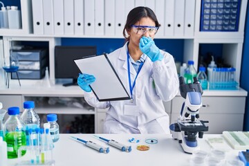 Hispanic young woman working at scientist laboratory smelling something stinky and disgusting,...