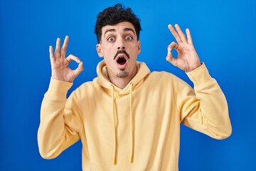 Naklejka premium Hispanic man standing over blue background looking surprised and shocked doing ok approval symbol with fingers. crazy expression