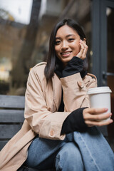 Cheerful asian woman in trench coat holding blurred paper cup and looking at camera near cafe.