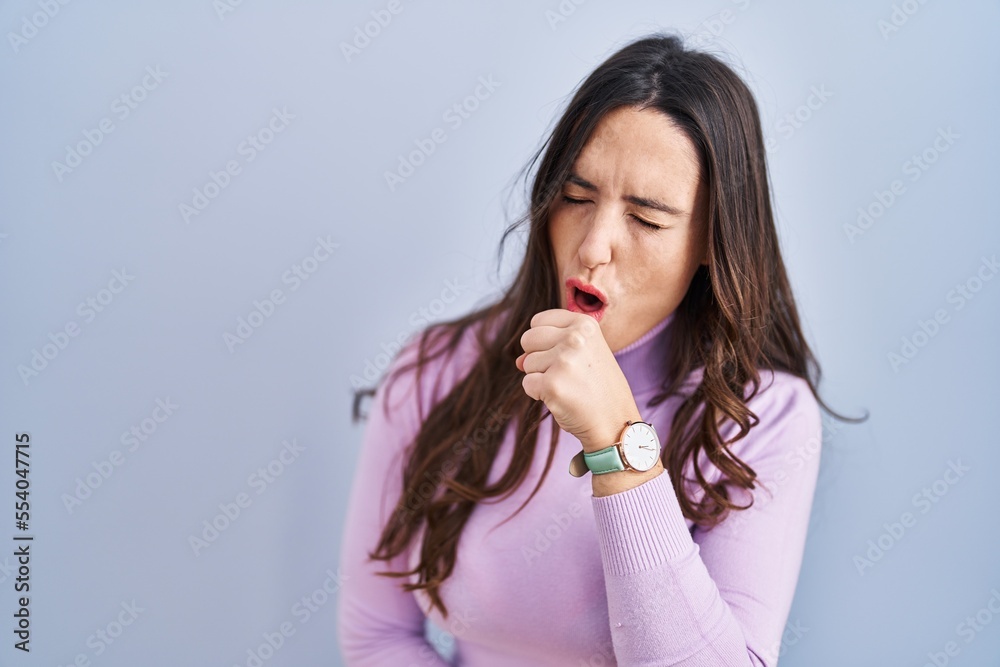 Poster young brunette woman standing over blue background feeling unwell and coughing as symptom for cold o