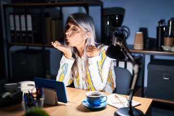 Middle age woman with grey hair working at the office at night looking at the camera blowing a kiss with hand on air being lovely and sexy. love expression.