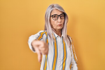 Middle age woman with grey hair standing over yellow background wearing glasses looking unhappy and...
