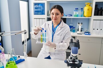 Young hispanic woman wearing scientist uniform holding test tubes at laboratory