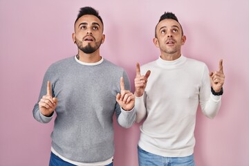 Homosexual couple standing over pink background amazed and surprised looking up and pointing with fingers and raised arms.