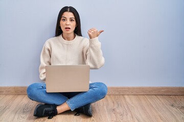 Young woman using laptop sitting on the floor at home surprised pointing with hand finger to the side, open mouth amazed expression.