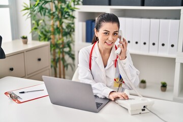 Young beautiful hispanic woman doctor using laptop talking on telephone at clinic