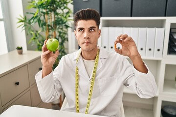 Young hispanic doctor man holding pills for fat loss making fish face with mouth and squinting eyes, crazy and comical.