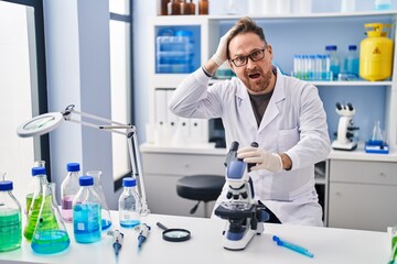Middle age caucasian man working at scientist laboratory stressed and frustrated with hand on head, surprised and angry face
