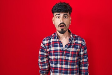 Young hispanic man with beard standing over red background afraid and shocked with surprise and amazed expression, fear and excited face.