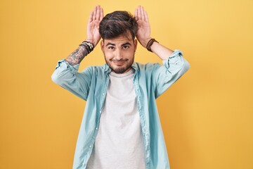 Young hispanic man with tattoos standing over yellow background doing bunny ears gesture with hands...
