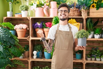 Young hispanic man with tattoos working at florist shop holding plant screaming proud, celebrating victory and success very excited with raised arm