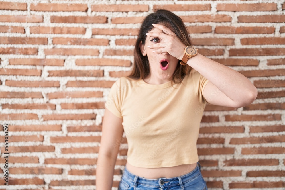 Wall mural Young brunette woman standing over bricks wall peeking in shock covering face and eyes with hand, looking through fingers with embarrassed expression.