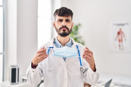 Young Hispanic Doctor Man With Beard Holding Safety Mask Skeptic And Nervous, Frowning Upset Because Of Problem. Negative Person.