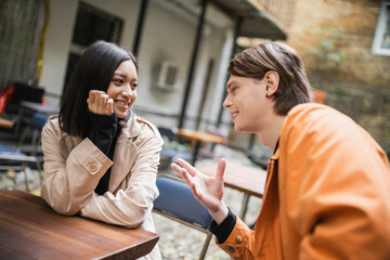 Smiling asian woman in trench coat looking at boyfriend talking in outdoor cafe.