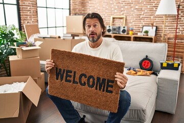 Handsome middle age man holding welcome doormat at new home in shock face, looking skeptical and...