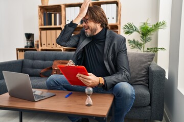 Handsome middle age man working at consultation office surprised with hand on head for mistake,...