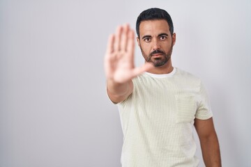 Fototapeta premium Hispanic man with beard standing over isolated background doing stop sing with palm of the hand. warning expression with negative and serious gesture on the face.