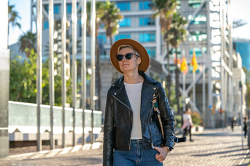 Obraz premium 30s woman with short blonde hair in leather jacket and trendy hat walking in city with modern building in Barcelona