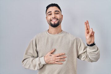 Young handsome man standing over isolated background smiling swearing with hand on chest and...