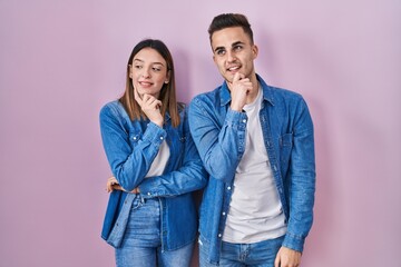 Young hispanic couple standing over pink background with hand on chin thinking about question, pensive expression. smiling and thoughtful face. doubt concept.