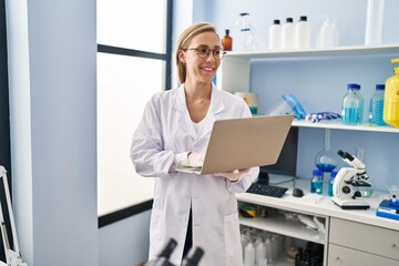 Young blonde woman scientist smiling confident using laptop at laboratory